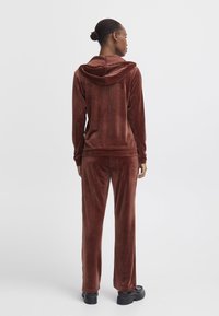 Brown velour tracksuit with a hooded top, featuring long sleeves and matching wide-leg pants. Smooth texture and relaxed fit. Black shoes.