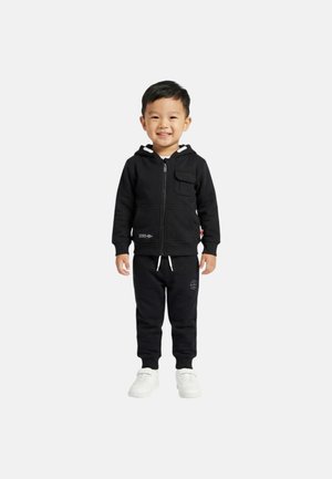 Jeune garçon souriant, portant un sweat à capuche noir zippé, un pantalon de survêtement noir assorti avec un logo, et des baskets blanches, debout devant un fond uni.