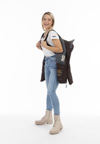 Femeie zâmbitoare cu păr blond, îmbrăcată cu un tricou alb, blugi deschiși la culoare și cizme bej, ținând o jachetă închisă pe umeri, pe un fundal alb.