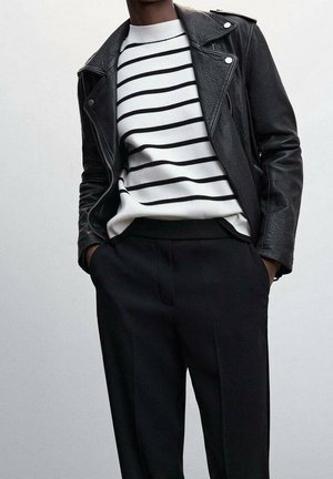 Une personne portant une veste en cuir noir sur un pull blanc avec des rayures horizontales noires et un pantalon noir, les mains dans les poches, sur un fond uni.