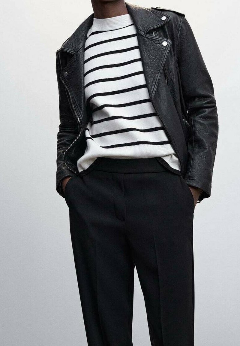 Person iført sort læderjakke over hvid sweater med sorte vandrette striber og sorte bukser, hænderne i lommerne, foran en ensfarvet baggrund.
