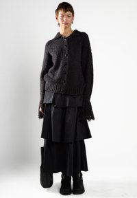 Kobieta w teksturowanym czarnym kardiganie zapinanym na guziki, z frędzlami na mankietach, warstwowej czarnej spódnicy, czarnych botkach i trzymająca czarną torebkę.