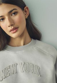 Világosszürke pulóver, amelyen a "NEW YORK" felirat hímzett, kiemelkedő, kissé sötétebb cérnával. Kerek nyakú, bordázott textúrával.