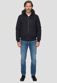 Schwarze Jacke mit Reißverschluss und Kapuze, mit rotem Innenfutter, seitlichen Taschen und einer Brusttasche. Kombiniert mit blauen Jeans und schwarzen Schuhen.