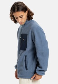 Világoskék fleece pulóver magas nyakú, sötétkék cipzáros mellzsebbel, texturált felülettel és oldalsó zsebekkel.