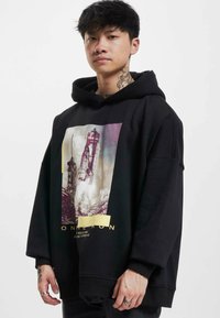 Svart överdimensionerad hoodie med ett grafiskt tryck av en raket och text. Den har en dragsko i huvan och ribbade muddar. Tyget verkar mjukt.