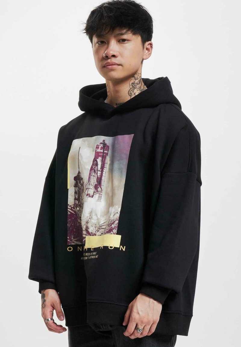 Svart överdimensionerad hoodie med ett grafiskt tryck av en raket och text. Den har en dragsko i huvan och ribbade muddar. Tyget verkar mjukt.