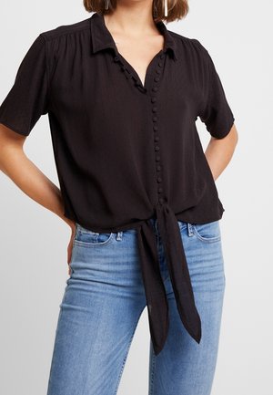 Kvinde iført en kortærmet sort knapbluse med knude foran kombineret med lyseblå jeans, stående med hænderne på hofterne.