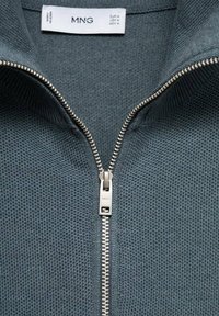 Knitted zip-up sweater in dark teal, features a textured pattern, a silver metal zipper, and a high collar. Brand label visible inside.
