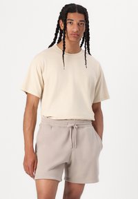 Camiseta de algodón beige de manga corta con un pequeño logo, combinada con pantalones cortos grises claros con cordón en la cintura que cuentan con bolsillos laterales y un ajuste relajado.