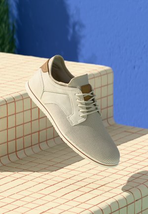Light beige sneaker with a mesh upper, brown leather accents, and flat laces, resting on a grid-patterned surface.