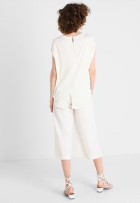 Krémový top s krátkými rukávy a zipem na zádech, spárovaný s širokými bílými kalhotami do culottes a bílými sandály na řemínky, s volným designem.