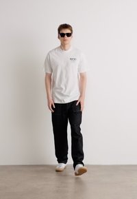 Hvit t-skjorte med logo, mørke denim bukser og hvite joggesko. Modell med svarte solbriller, står på en betonggulv mot en ensfarget bakgrunn.