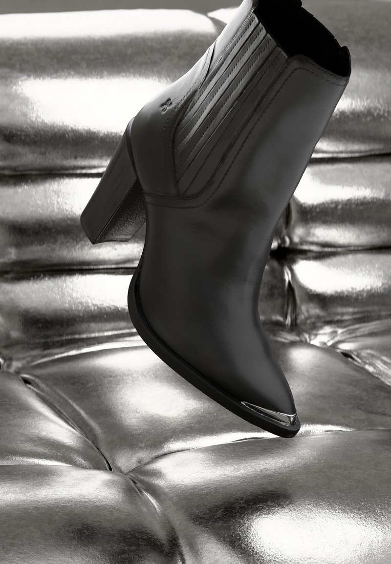 Bottine en cuir noir avec bout pointu et talon bloc. Présente une couture verticale et un accent métallique au niveau du bout. Placée au-dessus d'une surface brillante.