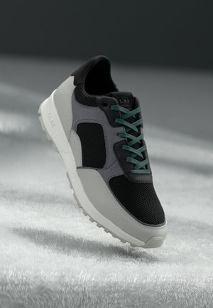 Szaro-czarny but sportowy z zielonymi sznurówkami i białą podeszwą unoszący się nad miękką, fakturowaną białą powierzchnią w delikatnym świetle.