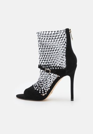 Czarna sandałka na wysokim obcasie z siateczkowym cholewką ozdobioną przezroczystymi kryształkami, otwartym palcem, paskiem wokół kostki i zamkiem błyskawicznym z tyłu.