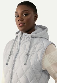 Ljust grå quiltad väst med huva, som har full dragkedja, snörningsdetaljer och silverfärgade metalldetaljer.