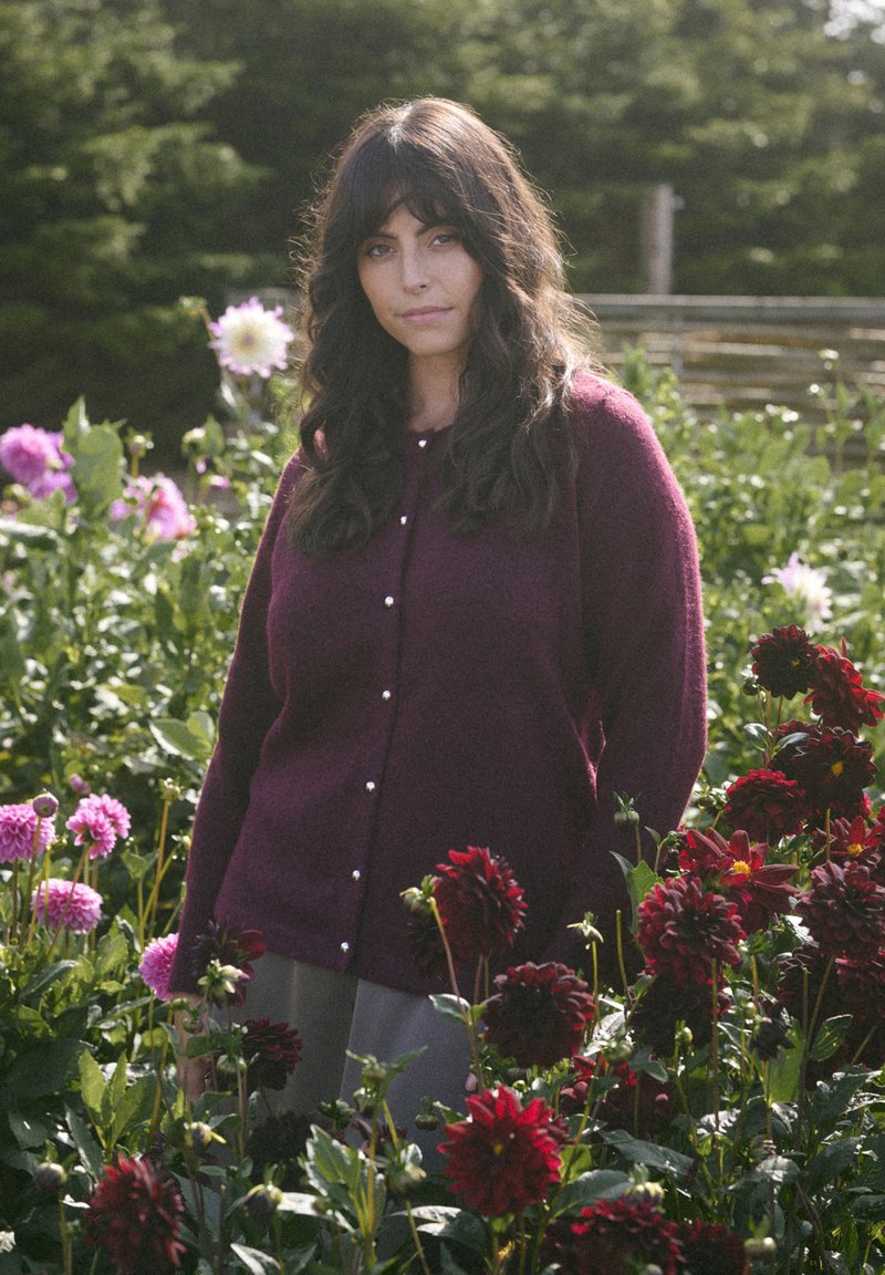 Mujer con cabello largo y oscuro, vestida con un cárdigan morado, se encuentra entre flores rojas y rosas en un jardín verde con fondo difuminado.