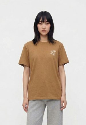 Una mujer está de pie mirando al frente con un fondo blanco liso, lleva una camiseta marrón con un pequeño logo en el pecho y pantalones grises.