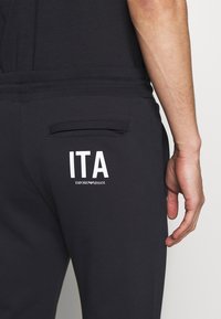Black sweatpants featuring a back pocket with a white "ITA" logo and "Emporio Armani" text, smooth texture, elastic waistband.