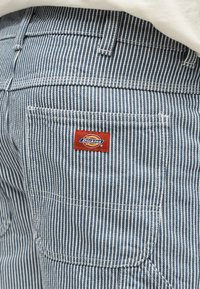 Τσέπη από παντελόνι με κάθετες ρίγες μπλε και λευκού χρώματος, με ραμμένη ετικέτα της μάρκας Dickies σε κόκκινο και κίτρινο χρώμα.