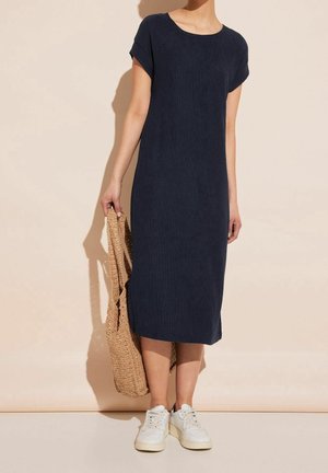 Femme portant une robe midi bleu marine à manches courtes, baskets blanches, tenant un grand sac fourre-tout tressé beige sur un fond beige.