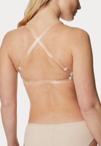 Beige beha met transparante criss-cross schouderbanden, voorzien van verstelbare hardware en soepele stof tegen een huidskleurover achtergrond.
