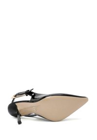 Zapato de tacón alto de charol negro con punta afilada, correa al tobillo y suela de un beige claro. Presenta un acento de hebilla metálica.