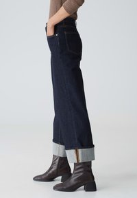 Donkerblauwe spijkerbroek met rechte pijpen, opgerolde zoom die grijze manchetten laat zien, gecombineerd met bruine enkellaarsjes. Stiksel zichtbaar langs de naden.