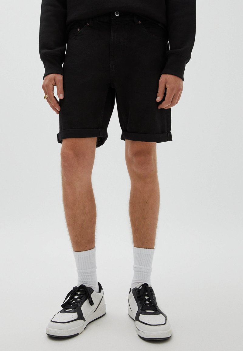 Partie inférieure du corps d'une personne portant un short en denim noir retroussé, des chaussettes blanches montantes, des baskets noires et blanches, et un haut à manches longues noir.
