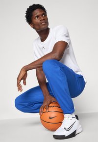 Niebieskie spodnie sportowe, biała koszulka z czarnym logo, białe i czarne sneakersy, przykucnięty z piłką koszykową, teksturowane powierzchnie i dopasowany krój.