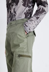 Pantalones cargo en mezcla de algodón verde oliva, con bolsillos laterales y un bolsillo con cremallera en el muslo, combinados con una camisa de manga larga gris con estampado.
