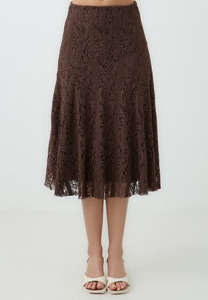 Jupe en dentelle marron avec un design évasé, des motifs en dentelle intricate et un ourlet à volants. L'association avec des sandales blanches à bout ouvert rehausse le look.