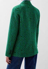 Groene blazer met textuur van zebra-strepen, op maat gemaakt ontwerp, gekraagde revers en knoopmanchetten. Gecombineerd met blauwe denim jeans.