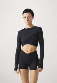 Crna dugosleeve cropped majica s uvrnutim prednjim dijelom i šorts s visokim strukom. Obje odjeće imaju glatku, pripijenu teksturu i minimalistički dizajn.