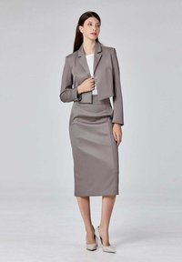 Costume gris ajusté avec une veste courte et une jupe cintrée, fabriqué en tissu lisse. Le mannequin porte des talons hauts de couleur claire.