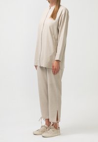 Béžový ležérní outfit skládající se z dlouhého zipového topu s dlouhým rukávem a odpovídajících zúžených kalhot s bočními rozparky, doplněný o světle barevné tenisky.