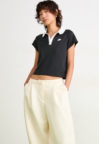 Ungdom med kort krøllete hår iført en svart Nike polo-shirt og løse kremfargede bukser, står med hendene i lommene mot en hvit vegg.