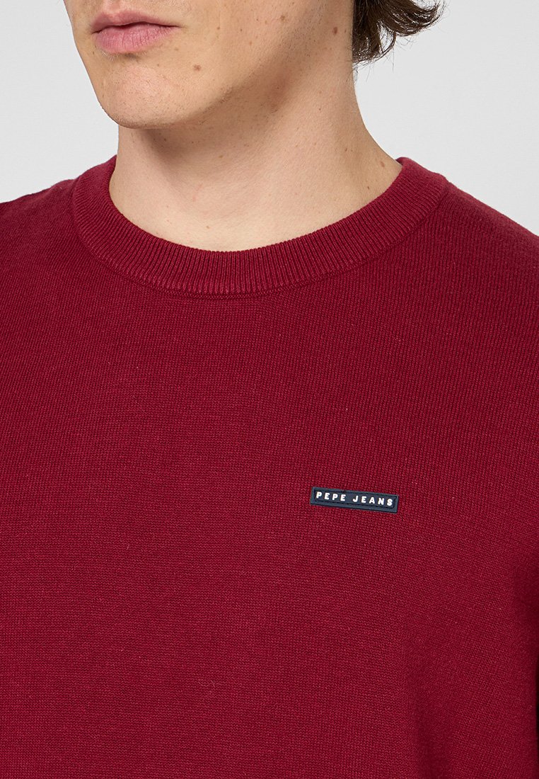 Pull en tricot rouge avec un col rond côtelé, arborant une petite étiquette noire "Pepe Jeans" sur le côté gauche de la poitrine. Texture douce.