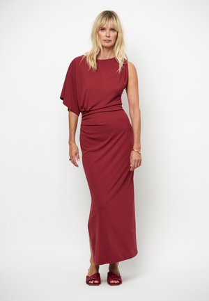 Femme blonde portant une robe maxi asymétrique bordeaux sans manches avec des chaussures ouvertes assorties, sur fond blanc.