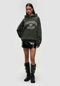 Hoodie oversize verde scuro con scritte grafiche bianche, abbinato a una mini gonna nera lucida e stivali alti neri con fibbie. Tessuti testurizzati e design sportivo.