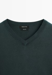 Maglione in cotone verde scuro a collo a V, con una texture liscia e bordi a coste. L'etichetta riporta "Massimo Dutti", indicando il marchio e il materiale.