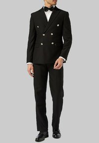 Costume noir croisé avec des boutons dorés, revers en pointe et pantalon assorti. Porté avec une chemise blanche et un nœud papillon noir. Chaussures noires.