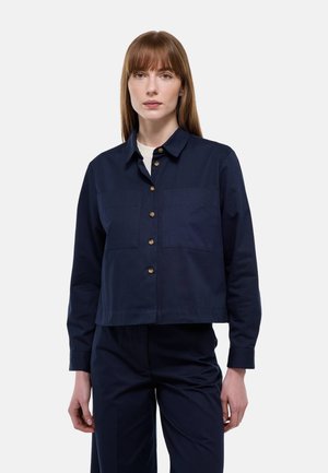 Femme aux cheveux roux lisses portant une chemise bleu marine à boutons avec deux poches poitrine et un pantalon assorti, sur un fond uni.