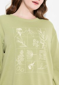 Felpa verde chiaro con un motivo a griglia di illustrazioni botaniche bianche e la parola "PURE" in grassetto. Materiale in cotone morbido con vestibilità comoda.
