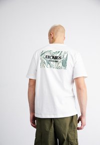 Camiseta blanca de algodón con un diseño botánico en verde y el texto "DICKIES" en negro y "SINCE 1922," sobre un fondo rectangular.