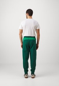 Gröna joggers med en kontrasterande grön midja, lös passform och elastiska muddar, i kombination med en enkel vit T-shirt. Synliga tatueringar på armen.
