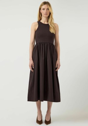 Kobieta o długich blond włosach, ubrana w czarną, bezrękawną sukienkę midi i brązowe, szpiczaste obcasy, stojąca na jednolitym jasnym tle.