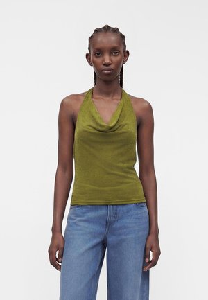 Femme aux cheveux tressés portant un haut dos-nu vert olive et un jean bleu, debout devant un fond gris clair uni.