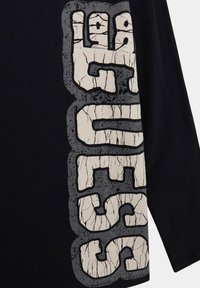 Tessuto nero con grandi lettere bianche screpolate contornate di grigio scuro che spelling "GUESS" in verticale lungo il lato.